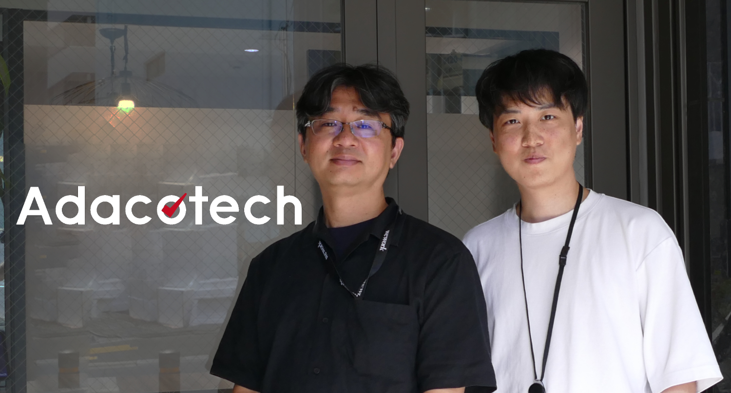 アダコテックのロゴを背にした株式会社アダコテック プロダクト開発部の伊部卓秀さんと瀧瀬航佑さん