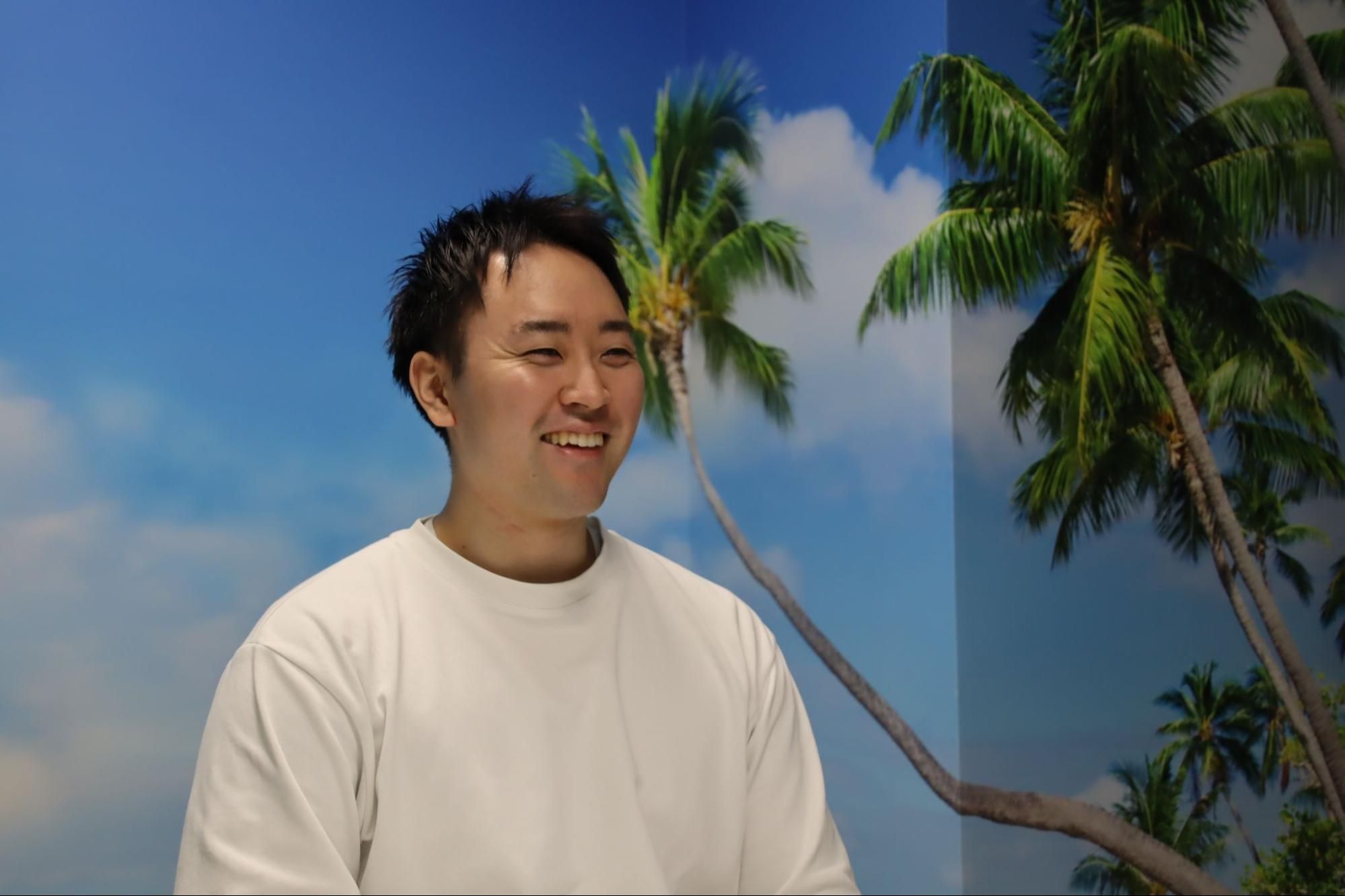 Young man in white t-shirt smiles against a blue sky with palm trees.